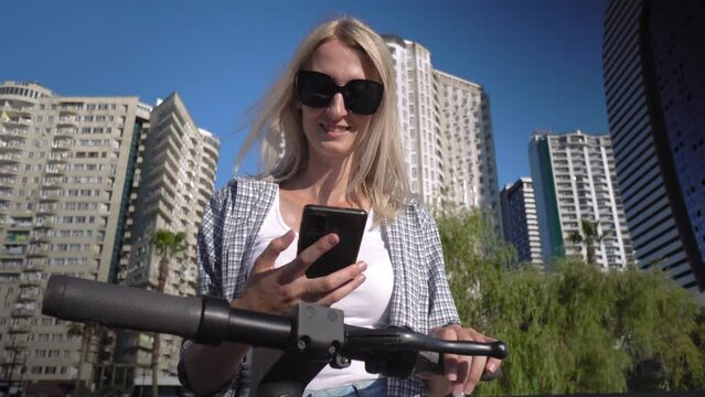 Young beautiful blonde woman in T-shirt and shirt uses app in her smartphone to rent eco-friendly electric scooter. Moving around city on hot day. eco-transport.