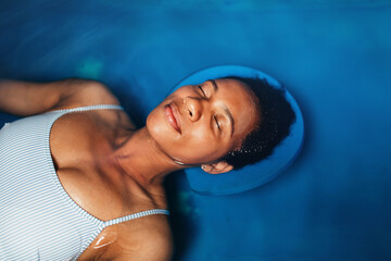 Beautiful middle-aged African American woman floating in tank filled with dense salt water used in meditation, therapy and alternative medicine.