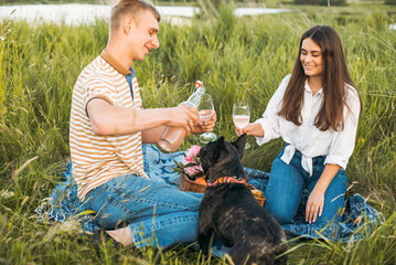 young happy couple having picnic with their french bulldog outdoors in the countryside during beautiful sunset. Romantic date near the lake