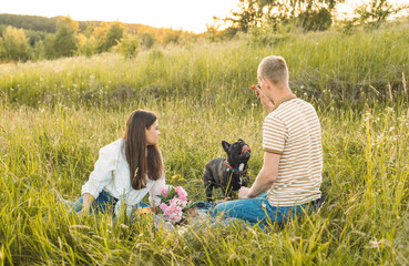 young happy couple having picnic with their french bulldog outdoors in the countryside during beautiful sunset. Romantic date near the lake