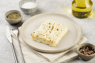 oven baked sheep cheese feta with herbs, oil and spices on round off-white plate, kitchen napkins, grey background