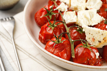 Roasted red tomatoes on branches with seasoned feta cheese on round off-white plate on kitchen napkins on grey background