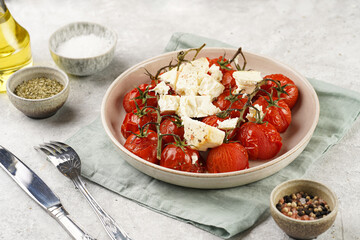 Roasted red tomatoes on branches with seasoned feta cheese on round off-white plate on kitchen napkins on grey background