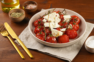 Roasted red tomatoes on branches with seasoned feta cheese on round off-white plate on kitchen napkins on wooden background