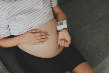 a pregnant woman sits on the couch and measures her blood pressure, her face is not visible