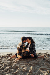 Young woman and man hugging while sitting together on the seashore with sea waves on the background at sunrise