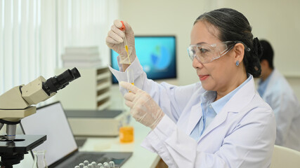 Professional middle aged female scientist in protective eyeglasses researching tube with reagents in laboratory