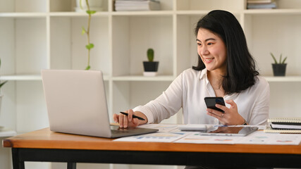 Pretty asian female employee holding mobile phone and using laptop on wooden office desk