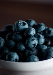 A handful of fresh blueberries in a white deep plate with handles on a wooden kitchen table in a dark modern interior. Topics of food, products, vitamins, health, cooking.
