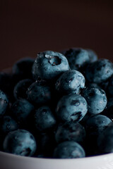A handful of fresh blueberries in a white deep plate with handles on a wooden kitchen table in a dark modern interior. Topics of food, products, vitamins, health, cooking.