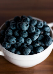 A handful of fresh blueberries in a white deep plate with handles on a wooden kitchen table in a dark modern interior. Topics of food, products, vitamins, health, cooking.
