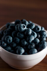 A handful of fresh blueberries in a white deep plate with handles on a wooden kitchen table in a dark modern interior. Topics of food, products, vitamins, health, cooking.