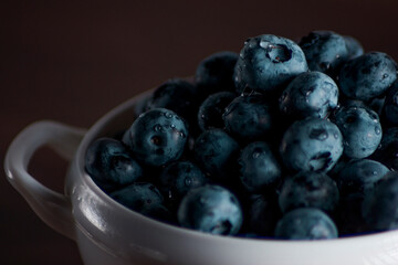 A handful of fresh blueberries in a white deep plate with handles on a wooden kitchen table in a dark modern interior. Topics of food, products, vitamins, health, cooking.