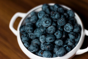 A handful of fresh blueberries in a white deep plate with handles on a wooden kitchen table in a dark modern interior. Topics of food, products, vitamins, health, cooking.