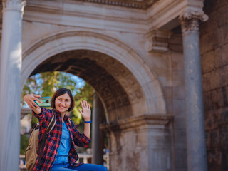 travel to Turkey. Happy asian female tourist traveller with backpack walks in old city. Woman takes selfie photos against backdrop of Hadrian's gate - popular attraction in old city of Antalya