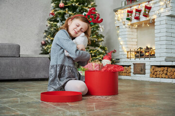 Happy kid girl opening Christmas gifts and hugs toy white bear. Child with presents in room. Merry Christmas and Happy Holidays