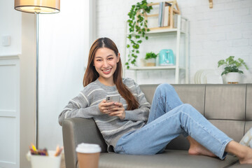Happy and cheerful woman is using a phone sitting on a sofa.Asian young woman looking mobile phone for shopping online at home.