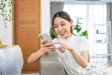 Happy and cheerful woman is using a phone sitting on a sofa.Asian young woman looking mobile phone for shopping online at home.