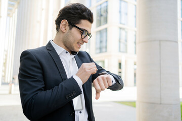 A businessman man with glasses looks at a smart watch uses an app, goes to work in a new office in a suit