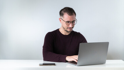 A man with glasses with a beard and mustache types on a laptop at his desk on a white background