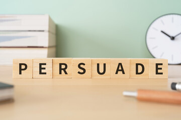 説得・納得のイメージ｜「PERSUADE」と書かれたブロックが置かれたデスク
