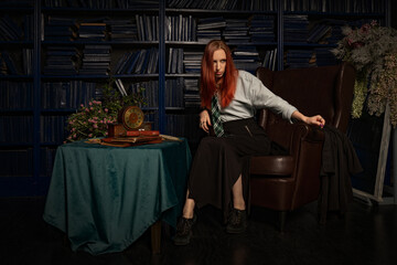 A young red-haired witch with a magic wand and a haughty expression sitting in an old library, witchcraft books and old clock on the table. She is dressed in a white shirt, black skirt and green tie