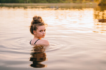 Teenager cheerful girl is bathing and looking at the camera over his shoulder merrily in a picturesque forest lake.
