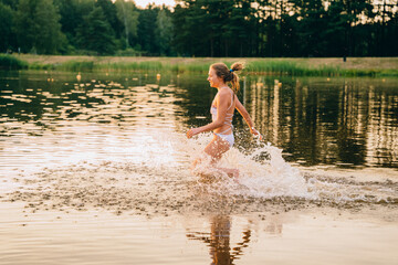Feeling freedom and fresh air. Happy active teenager girl in swimsiut running on the lake or river on a hot summer day at sunset. Summer cump, happy childhood, active lifestyle concept.