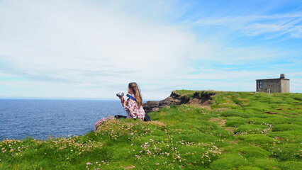 Downpatrick Head, County Mayo, Ireland - 07.07.22:

Young lady sitting at the marshy cliff edge with an analogue camera, overlooking the vast Atlantic Ocean.