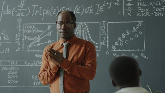 African American professor standing in front of blackboard with math formulas and explaining lesson to students in college