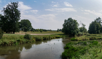 Panorama rzeki Osobłogi w obszarach polnych, w tle zarośla i nieliczne drzewa w porze letniej na tle nieznacznie pochmurnego nieba - obrazy, fototapety, plakaty