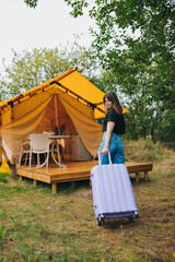 Fototapeta na wymiar Rear view of woman traveler with luggage moving towards bedroom of cozy glamping house. Luxury camping tent for outdoor summer holiday and vacation. Lifestyle concept