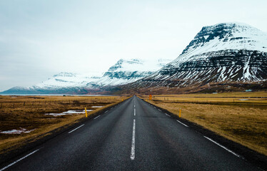 Panoramic winter photo of road leading along coast of lake to volcanic mountains. High rocky peaks covered with snow layer mirroring on water surface. Driver's point of view on Ring road, Iceland.

