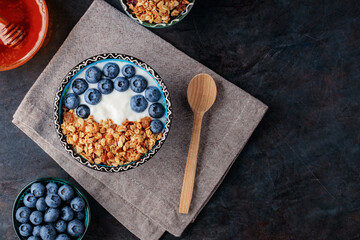 Granola, yogurt, blueberries and honey on a dark background. Bowl with yogurt, baked granola and berries on linen napkin. Top view. Copy space