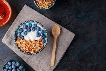 Granola, yogurt, blueberries and honey on a dark background. Bowl with yogurt, baked granola and berries on linen napkin. Top view. Copy space