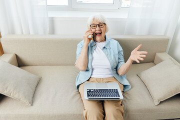 a joyful woman is sitting relaxed on a cozy sofa talking on a smartphone, holding a laptop on her lap while working from home, and gesticulating with her hand