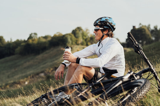 Professional Male Cyclist Drinking Water from Bottle, Man Sitting Near His Bicycle During His Journey Outdoors in Countryside