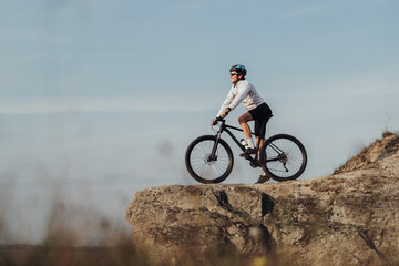 Equipped Professional Male Cyclist Standing with Mountain Bike on Edge of the Rock, Sky on the Background and Copy Space