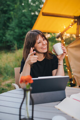 Happy Woman freelancer using laptop and drink coffee on a cozy glamping tent in a sunny day. Luxury camping tent for outdoor summer holiday and vacation. Lifestyle concept