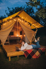 Obraz na płótnie Canvas Happy Woman freelancer using a laptop on a cozy glamping tent in a summer evening. Luxury camping tent for outdoor holiday and vacation. Lifestyle concept
