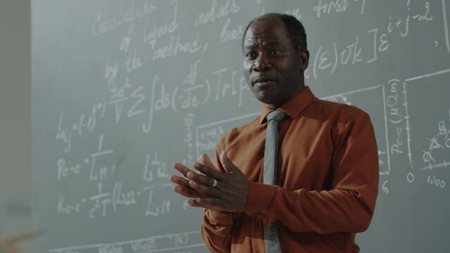Waist up shot of African American professor pointing at blackboard with formulas on it and explaining algebra during class time in university