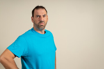 Serious and angry bearded man in blue t-shirt posing isolated on beige background, studio portrait. People lifestyle concept. Mock up copy space. Standing with your arms akimbo on your waist blinking
