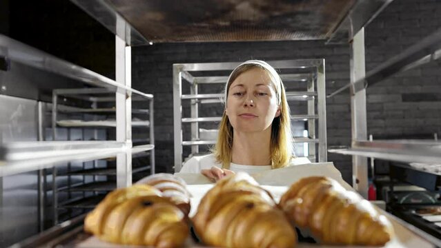Caucasian beautiful woman in hat and uniform working in bakehouse and putting croissants on shelf. Female baker making fresh pastries in kitchen of bakeshop in morning. Confectionery producing concept