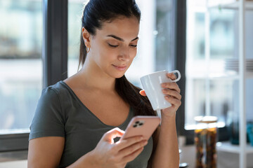 Beautiful young woman using her mobile phone while drinking a cup of coffee in the kitchen at home.