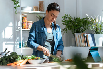 Healthy young woman cutting fresh vegetables and using digital tablet to recipes in the kitchen at home.