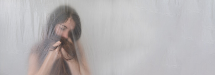 blurred, fuzzy wide panoramic banner portrait of depressed sad young woman with long hair puts head in hands, behind transparent plastic foil film