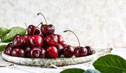Juicy ripe cherries on a ceramic plate. A plate of berries on a linen napkin. Red cherry berries on a light background with branches of greenery. Selective focus. The concept of healthy eating.