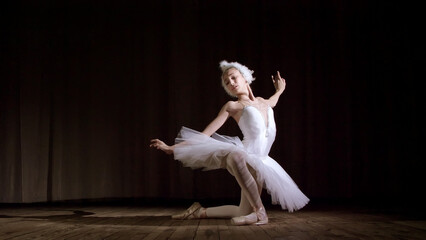 in rays of spotlight, on the stage of the old theater hall. Young ballerina in suit of white swan...