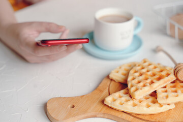 Person writes messages on smartphone and breakfast. Belgian waffles on desk, cup of coffee and honey stick on white table. Morning romantic pastime. Online communication over lunch. Business lady.