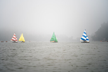 four colorful sailboats lost in the haze fog with smaller pedal row boats floating around on the...